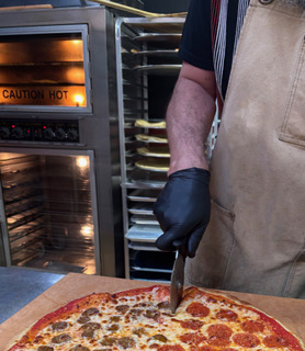 A cook slicing a pepperoni pizza next to a hot oven