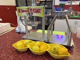 A fresh lemonade juicer with a tray of lemons and a We Squeeze to Please sign